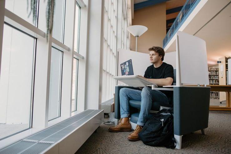 在立博体育登录的图书馆里，一名男生拿着笔记本电脑坐在椅子上望着窗外